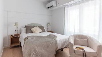A modern hotel room with a neatly made bed, a bedside table with a lamp, a chair, and a large window with sheer curtains.