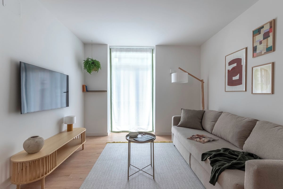 A modern living room with a sofa, a coffee table, a TV mounted on the wall, and a large window with curtains.