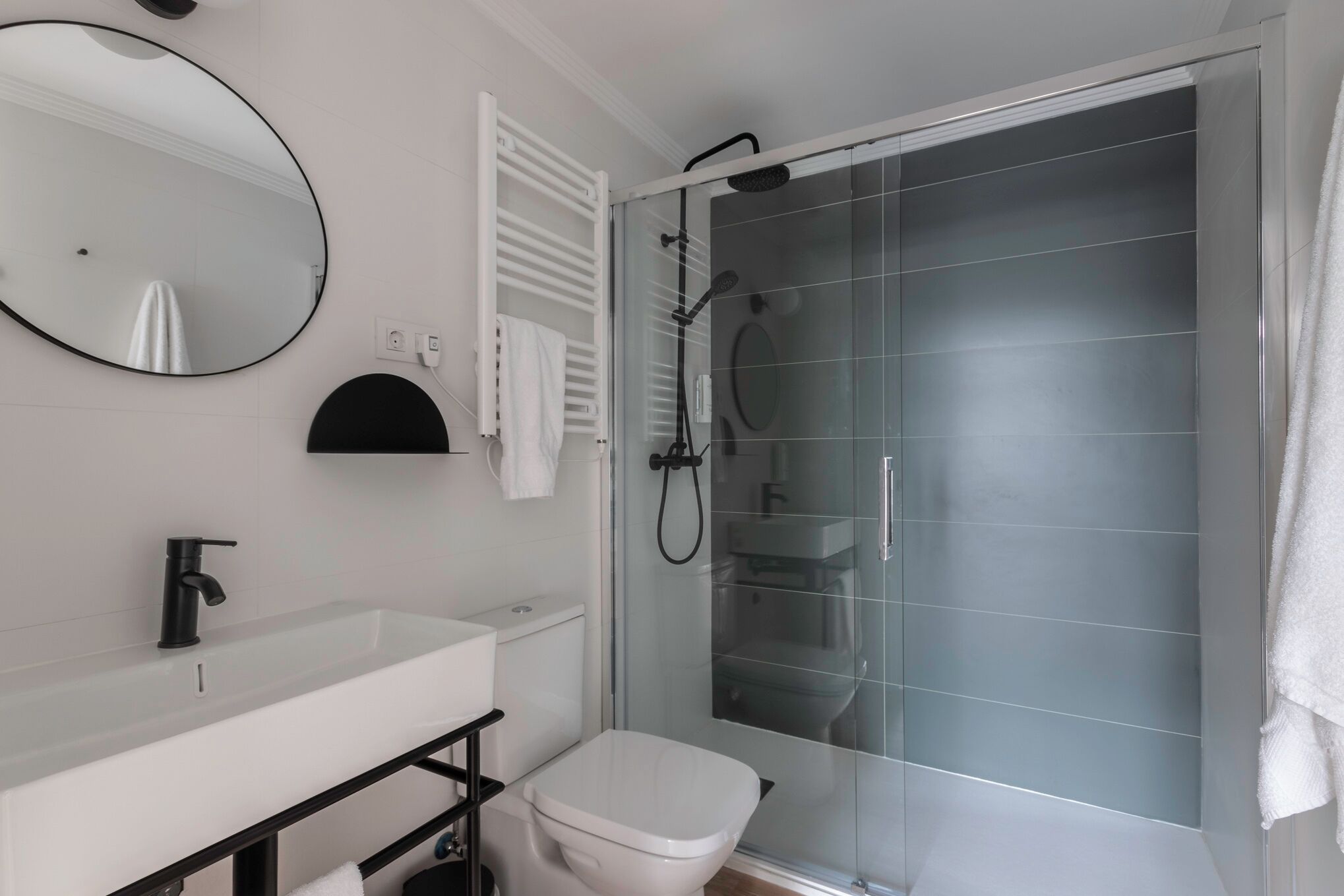 A modern bathroom with a white sink, black faucet, a round mirror, a toilet, and a glass-enclosed shower area.