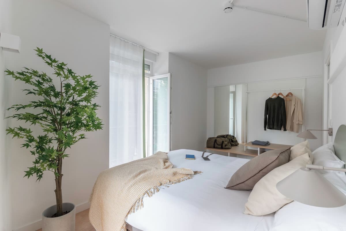 A hotel room with a bed, a bedside table, a lamp, a mirror, a wardrobe, and a potted plant.