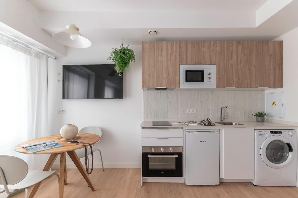 A modern kitchen with a built-in oven, microwave, and washing machine, a dining area with a table and chairs, and a large flat-screen TV mounted on the wall.