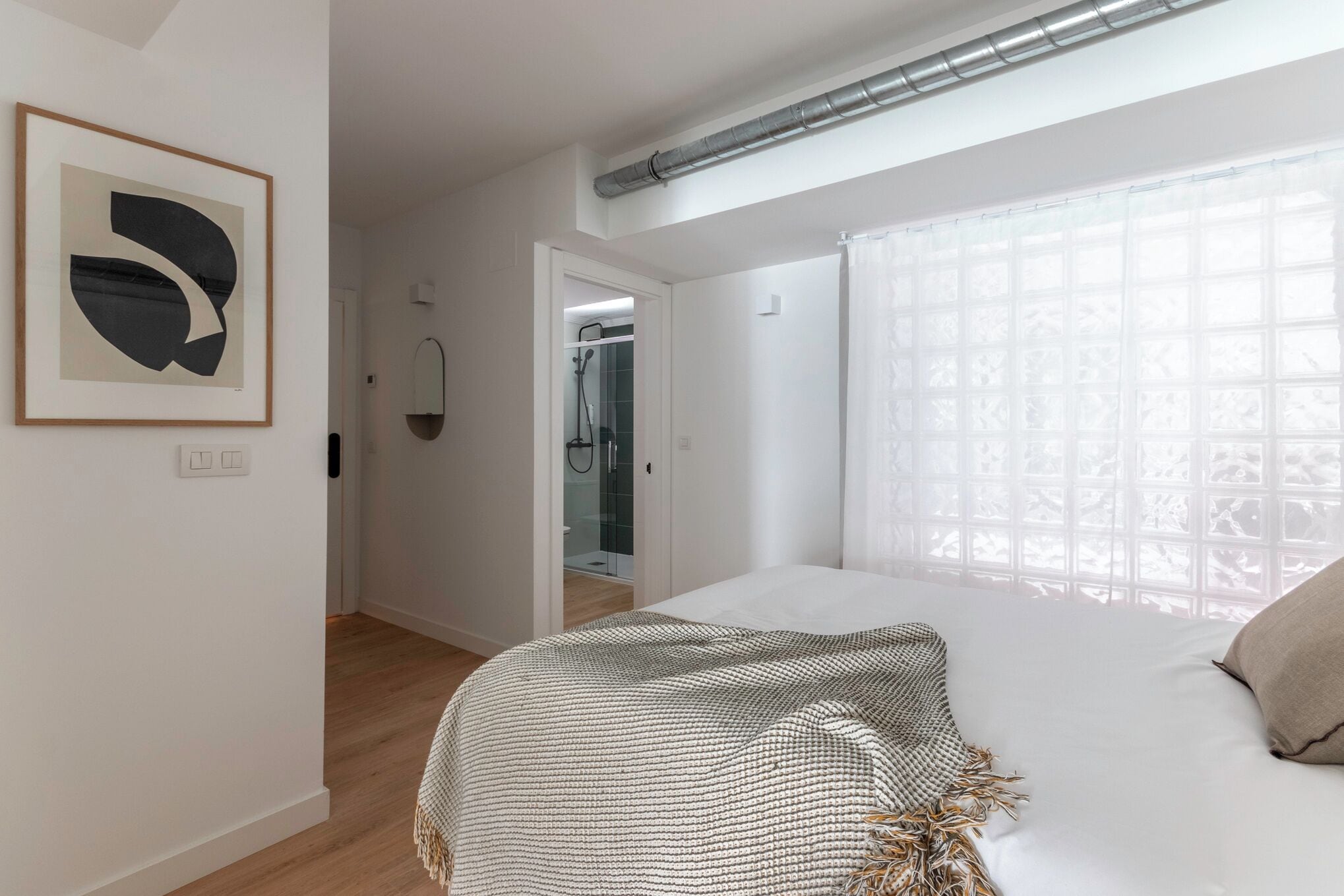 Un dormitorio con cama, una obra de arte abstracta en marco, zona de ducha y ventana con diseño de cuadrícula.