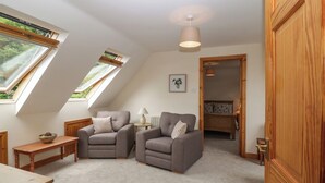 Een gezellige kamer met twee grijze fauteuils, een klein houten tafeltje en een lamp. Er zijn dakramen en een deur naar een andere kamer.