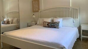 Un lit blanc avec une tête de lit, une table de chevet avec une lampe et un miroir au mur.