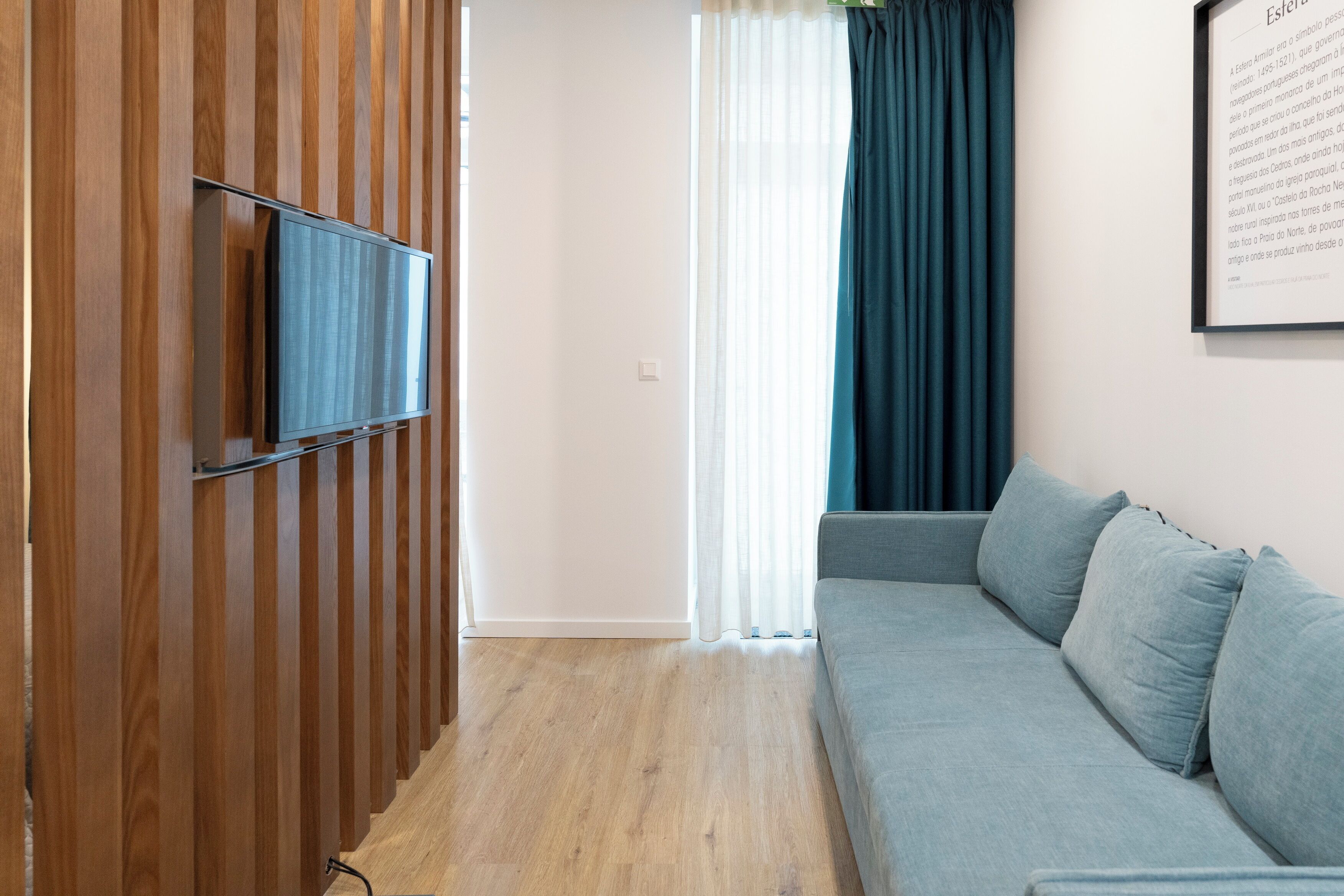 Ett modernt rum med en väggpanel i trä, en platt-TV som är monterad på väggen, en ljusblå soffa och ett fönster med tunna, vita gardiner och en mörkblå gardinkappa.