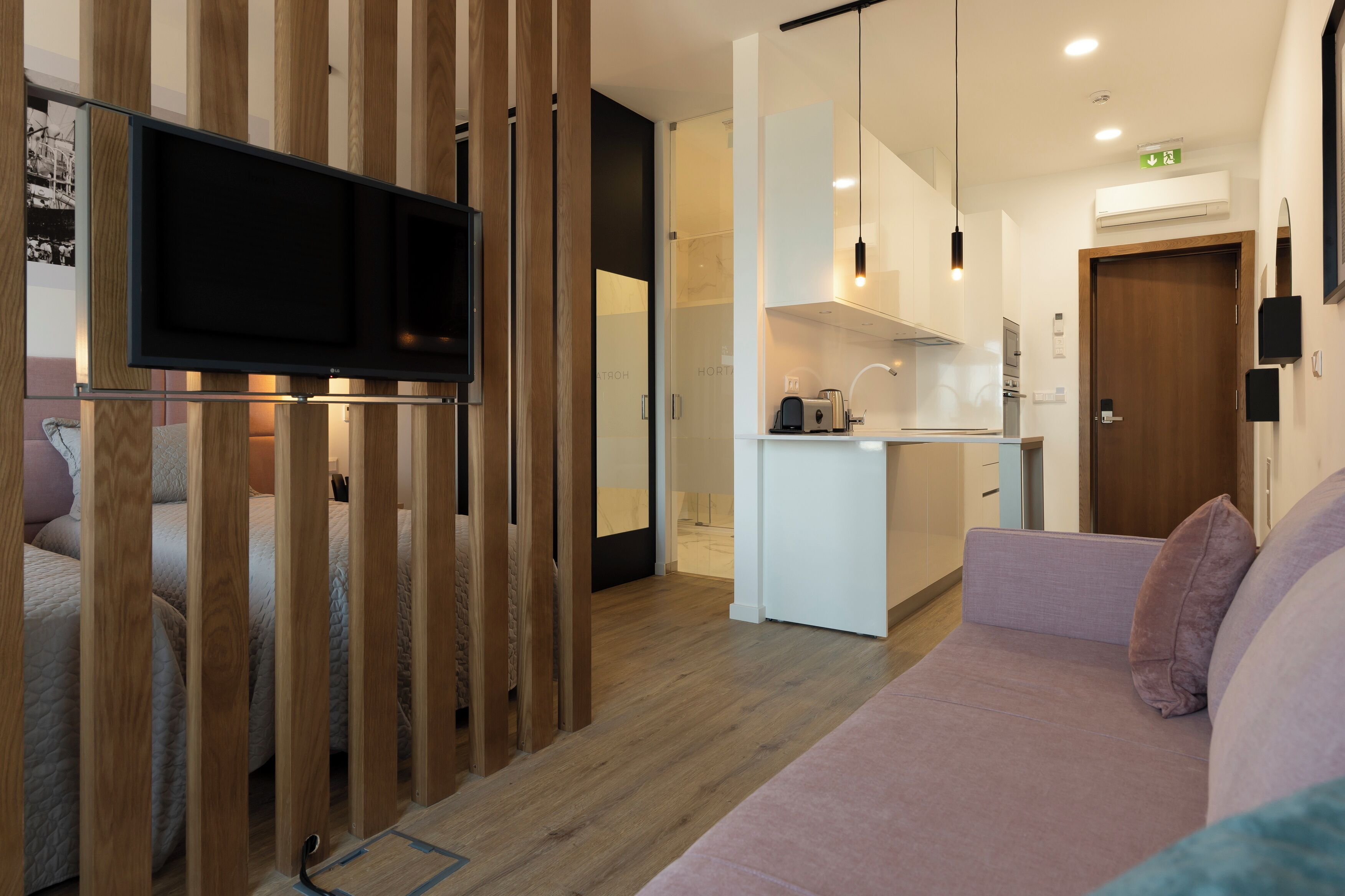 A modern living room with a sofa, a TV mounted on a wooden partition, and a kitchenette in the background.