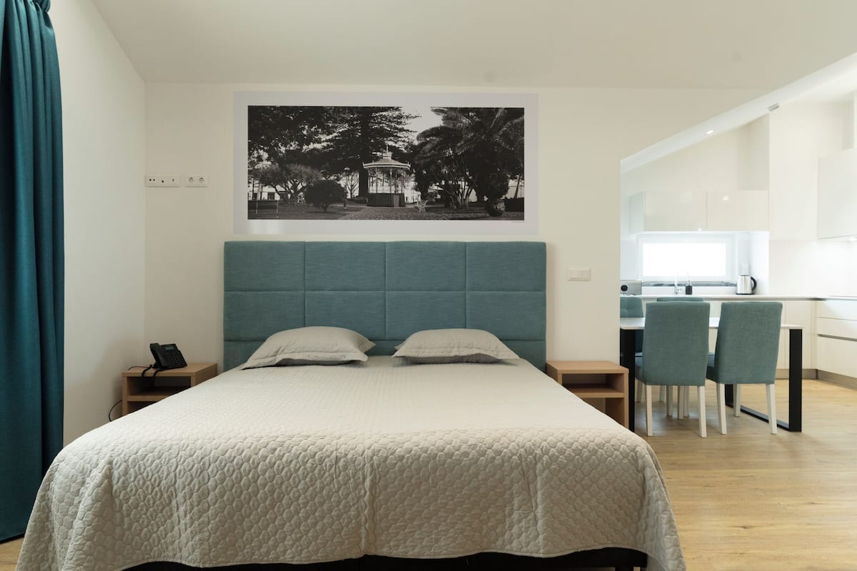 A modern bedroom with a large bed, a headboard, a bedside table with a phone, and a view of a dining area with chairs and a table.