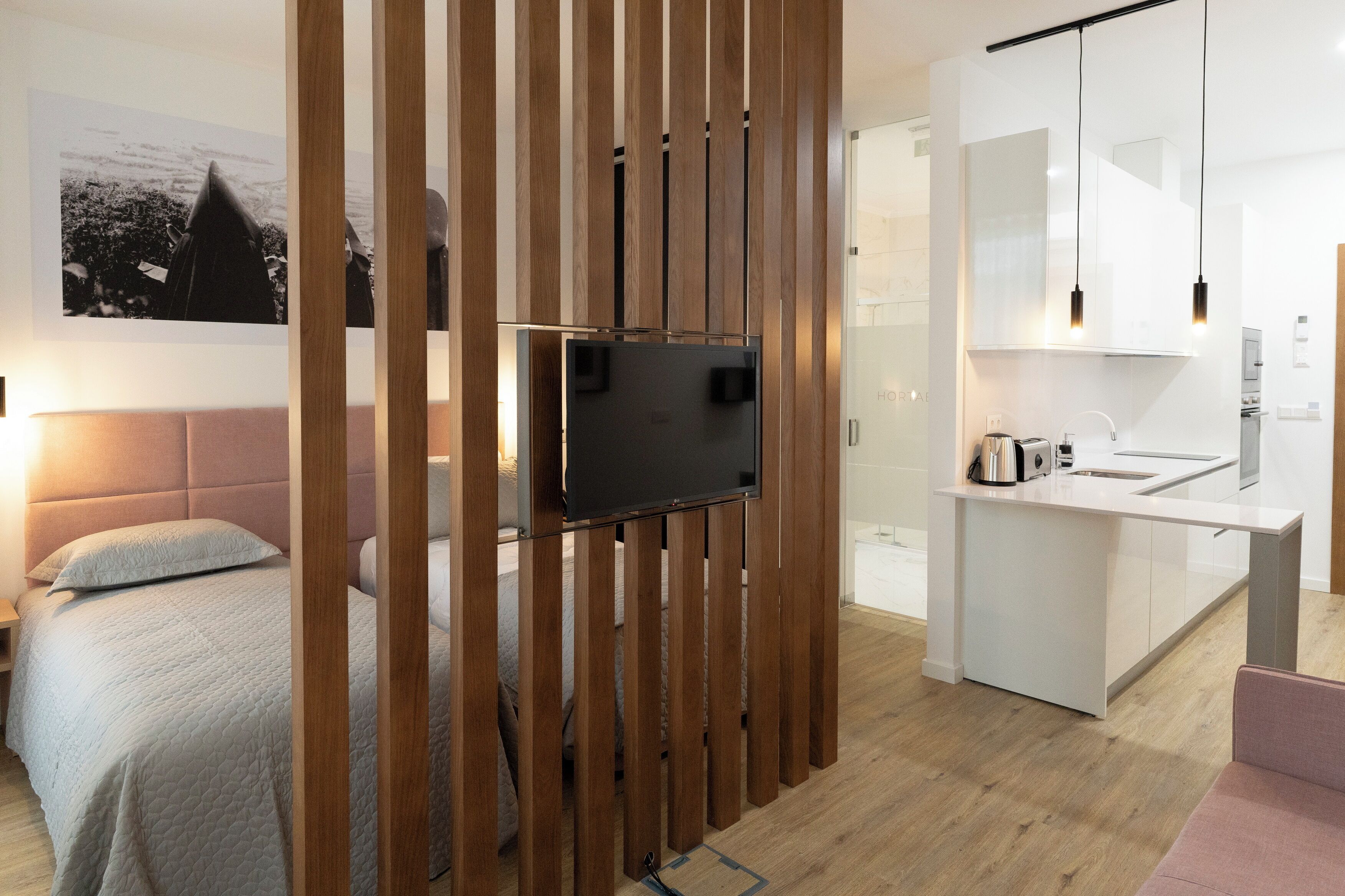 Una habitación moderna con una cama, un televisor montado en una pared de madera, una kitchenette con un fregadero y armarios, y un ventanal grande.