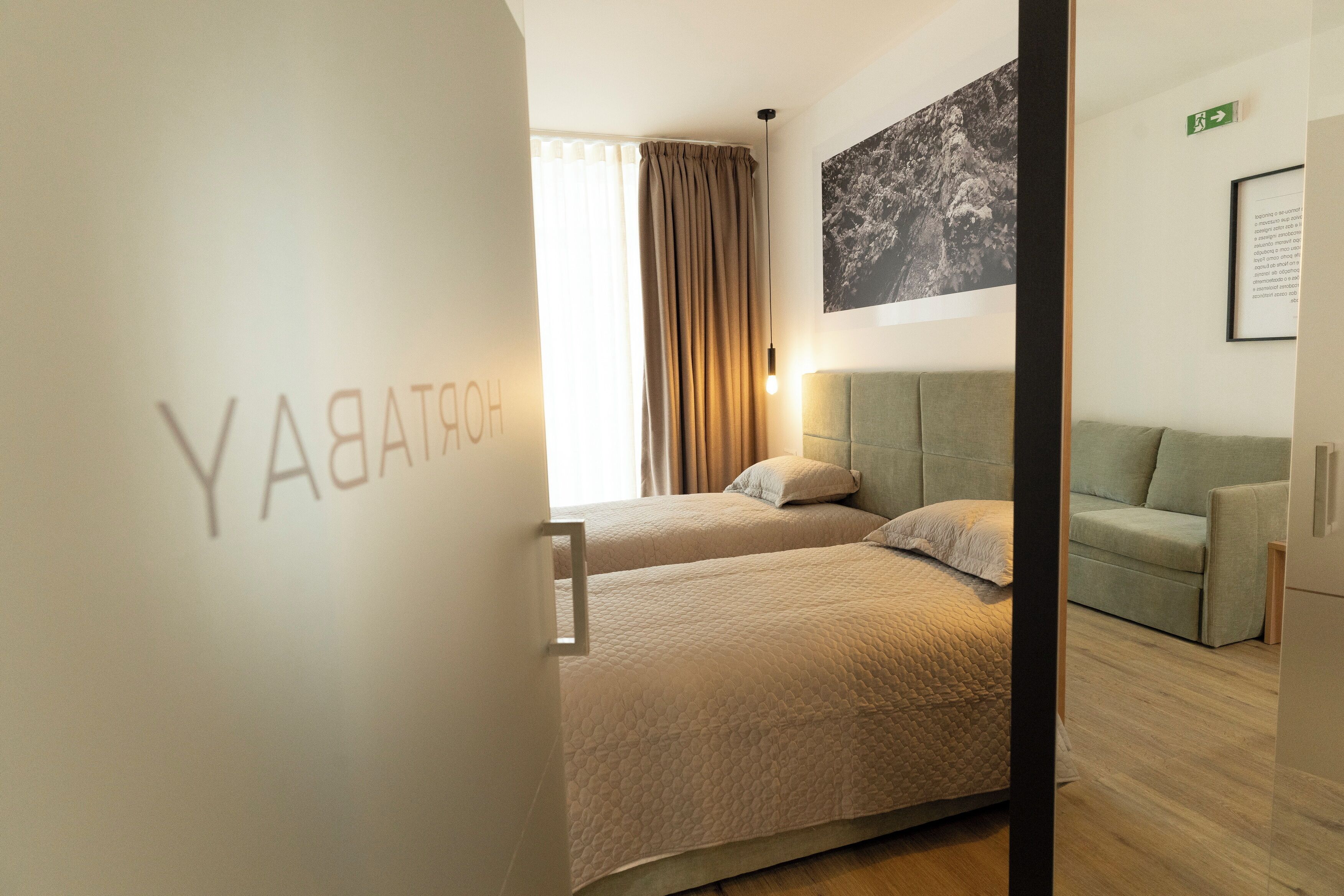Hotellihuone, jossa on kaksi sänkyä, vihreä sohva, suuri ikkuna verhoilla sekä seinälle kiinnitetty taideteos.