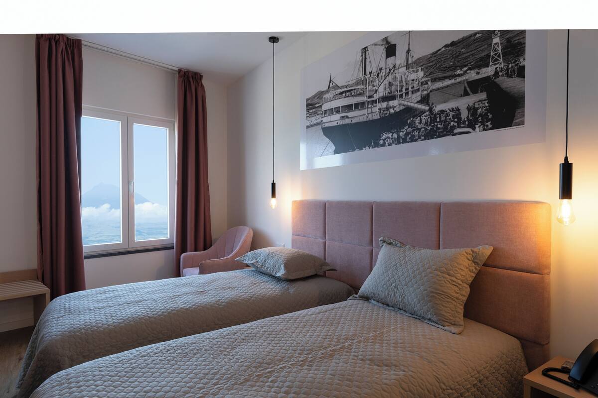 A hotel room with two beds, a chair, a nightstand, a lamp, and a large window with curtains.