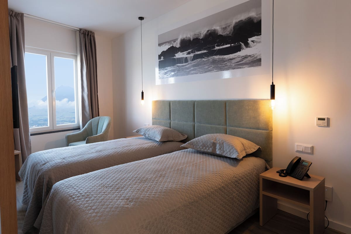 A hotel room with two beds, a chair, a bedside table with a phone, and a large window with curtains.