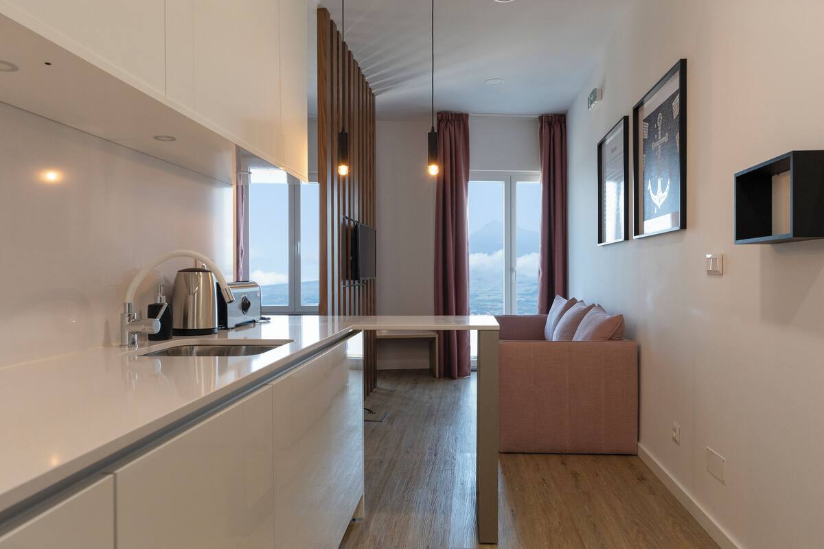 A modern kitchen with a built-in oven, a coffee machine, and a sink. There is a dining area with a table and chairs, and a sofa with cushions. The room has large windows with curtains, and artwork on the walls.