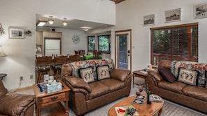 A living room with a brown leather sofa, a wooden coffee table, and a dining area with a green table and chairs. - Woodlands Way Chalet- A cozy get-away in Cloudcroft NM.
 (Cloudcroft)