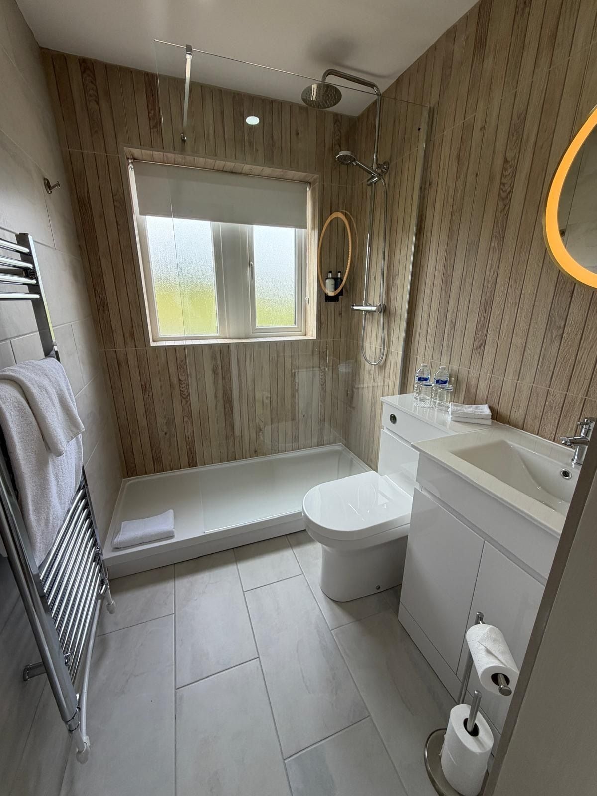 A modern bathroom with a wooden panel wall, a window, a shower, a toilet, and a sink.