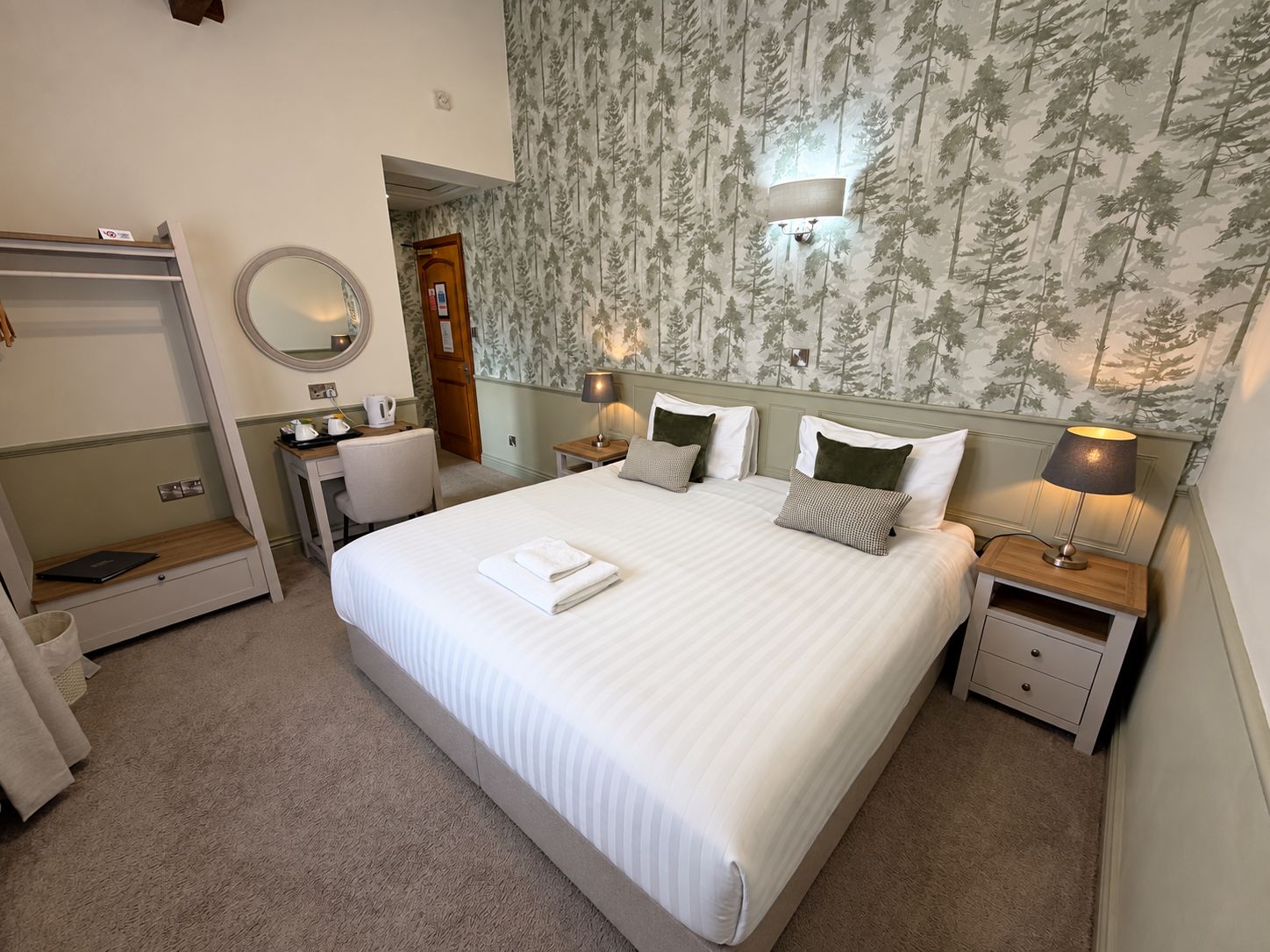 A hotel room with a large bed, bedside tables, a desk, a chair, a wardrobe, and a mirror.