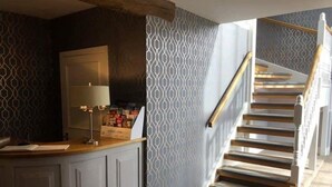 Una escalera de madera con barandilla blanca, que lleva a un rellano con un revestimiento de pared decorativo y una encimera con una lámpara y un atril para revistas.