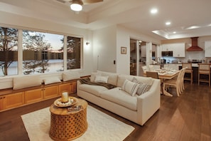 A modern living room with a large window, a sofa, a dining area, and a kitchen.