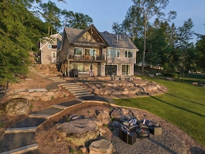 A two-story house with a stone staircase leading to a deck, surrounded by a well-maintained lawn and large rocks.