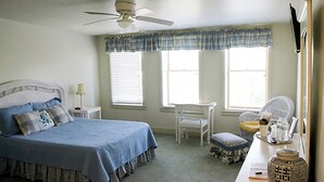 Une chambre à coucher comprenant un lit, un bureau, une chaise, un ventilateur de plafond et une fenêtre avec des stores.