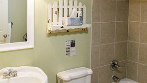 Une salle de bain avec un lavabo, des toilettes et une baignoire. Au-dessus des toilettes, on trouve une étagère décorative en forme de clôture blanche.