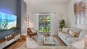 Un salón moderno con televisor de pantalla plana, un sofá, una mesa de centro y vistas a un espacio exterior con plantas a través de un ventanal.
