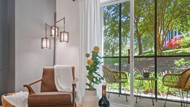 Una habitación con una silla marrón, un jarrón blanco con una botella, cestas de mimbre y vistas al verdor exterior.