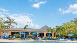 A pool area with lounge chairs and umbrellas, surrounded by palm trees and a building with a grey roof.