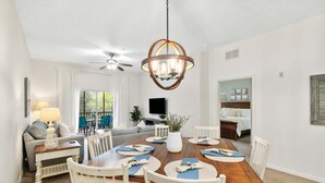 A dining area with a wooden table, white chairs, a chandelier, and a television in the background.