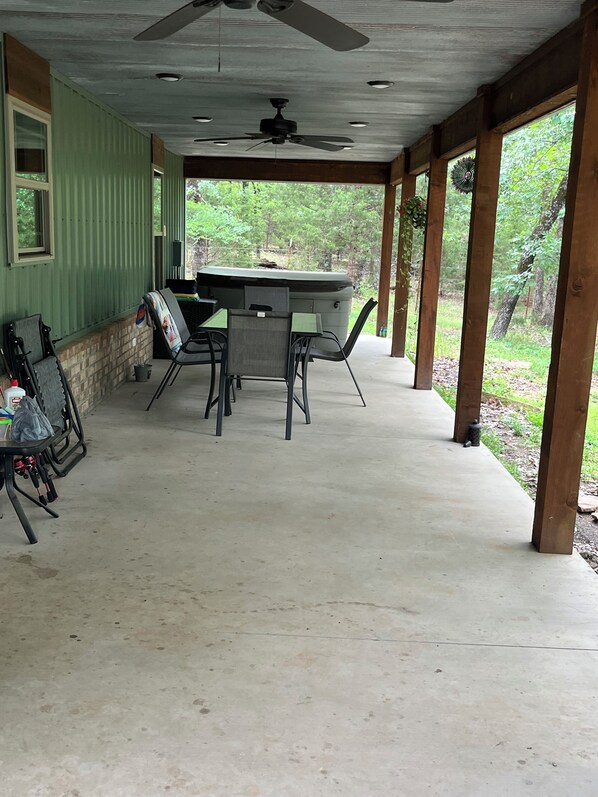 Een overdekt terras buiten met een tafel, stoelen, een plafondventilator en uitzicht op de omliggende begroeiing.