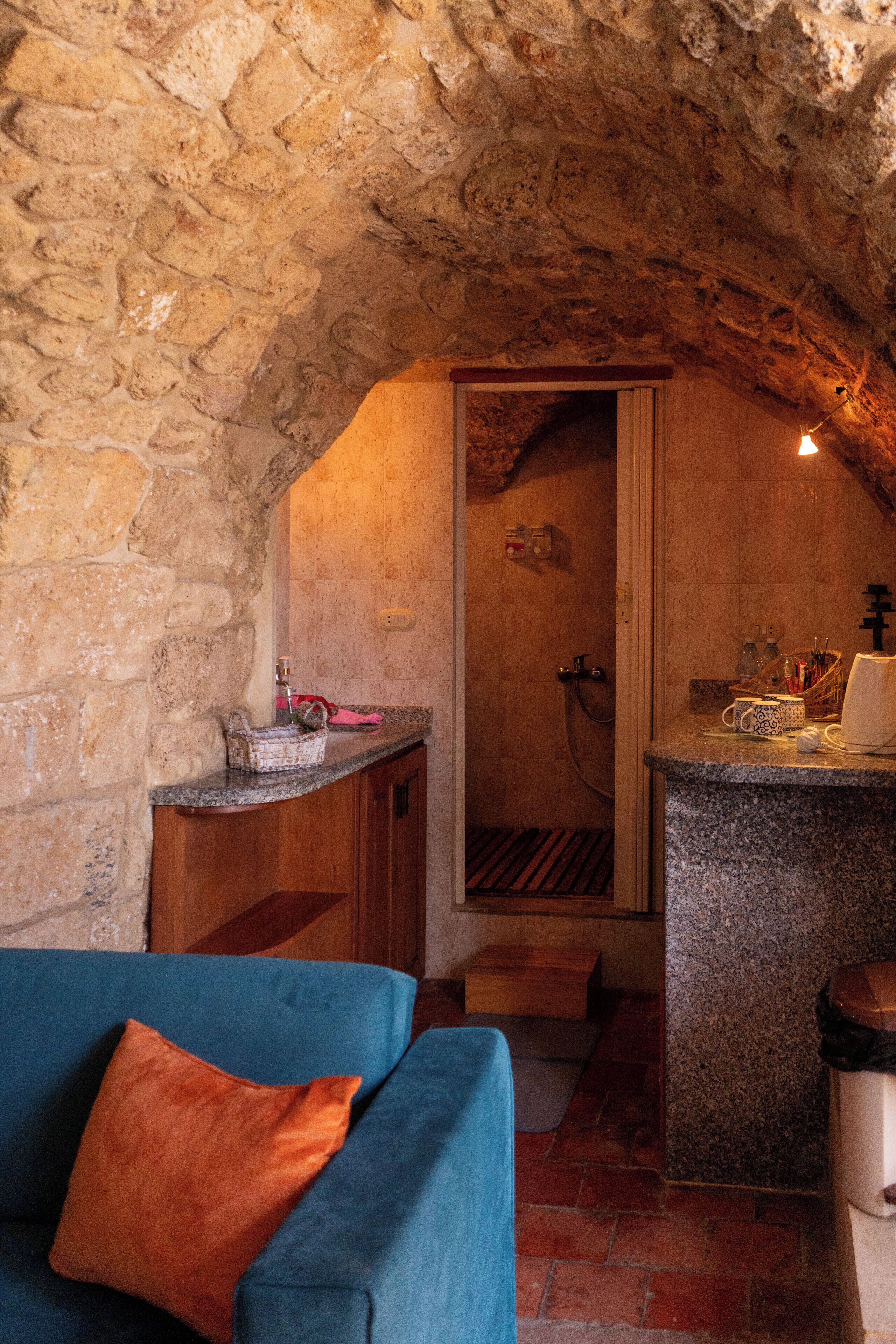 A stone-walled room with a wooden bench, a blue sofa with an orange cushion, and a small kitchen area with a sink and countertop.
