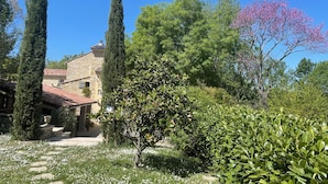 Un edificio de piedra con techo de tejas rojas, rodeado de exuberante vegetación y altos cipreses.