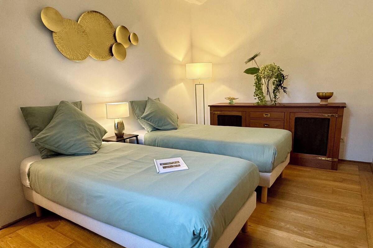 A hotel room with two beds, a nightstand with a lamp, a wooden dresser, and a wall-mounted decorative piece.