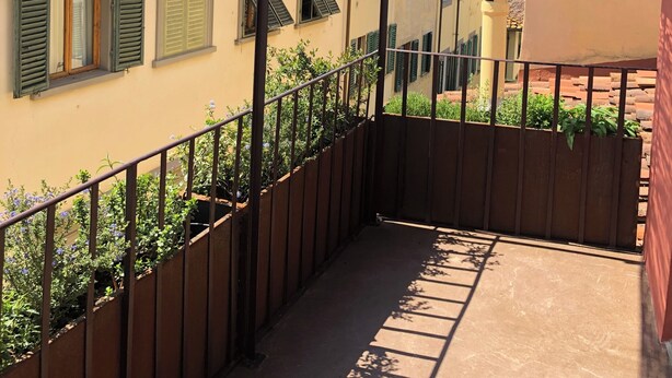 A balcony with a metal railing, a tiled floor, and a view of neighboring buildings with red-tiled roofs and green shutters. - Florence from above (Firenze)