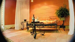 Um pátio com um banco de metal, uma mesa com bule e xícara, e um vaso com planta.