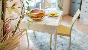 Een eethoek met een tafel gedekt voor twee personen, een schaal met snacks en een bord met salade. Er is een stoel en een klein tafeltje met een decoratief object. De kamer heeft uitzicht op de zee en bergen door grote ramen.