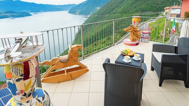 Een balkon met rieten meubilair, een houten paardbeeld en uitzicht op een meer en bergen.