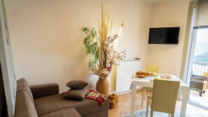 Een woonkamer met een bruine bank, een eettafel met stoelen, een vaas met planten en een televisie aan de muur.
