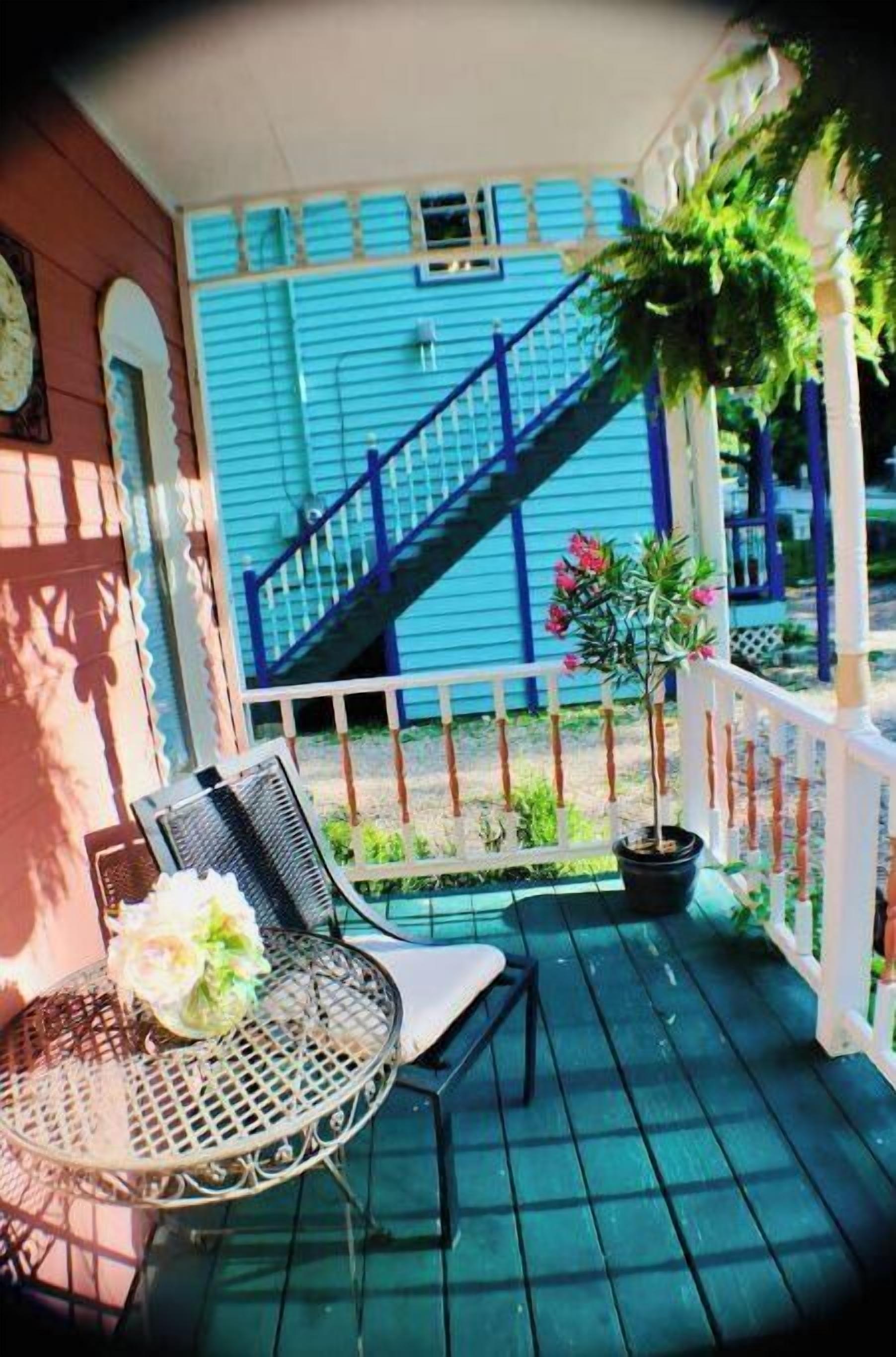 Un porche avec une chaise en métal et une petite table, une plante en pot et un escalier menant à un bâtiment bleu.