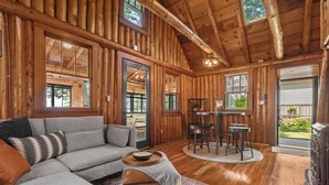 A log cabin interior with a gray sectional sofa, a wooden dining table with chairs, and a large window. - Lakefront Log Cabin-Stunning Lake Views, Kayaks, Steps to Beach & dt Lake City! (Lake City)
