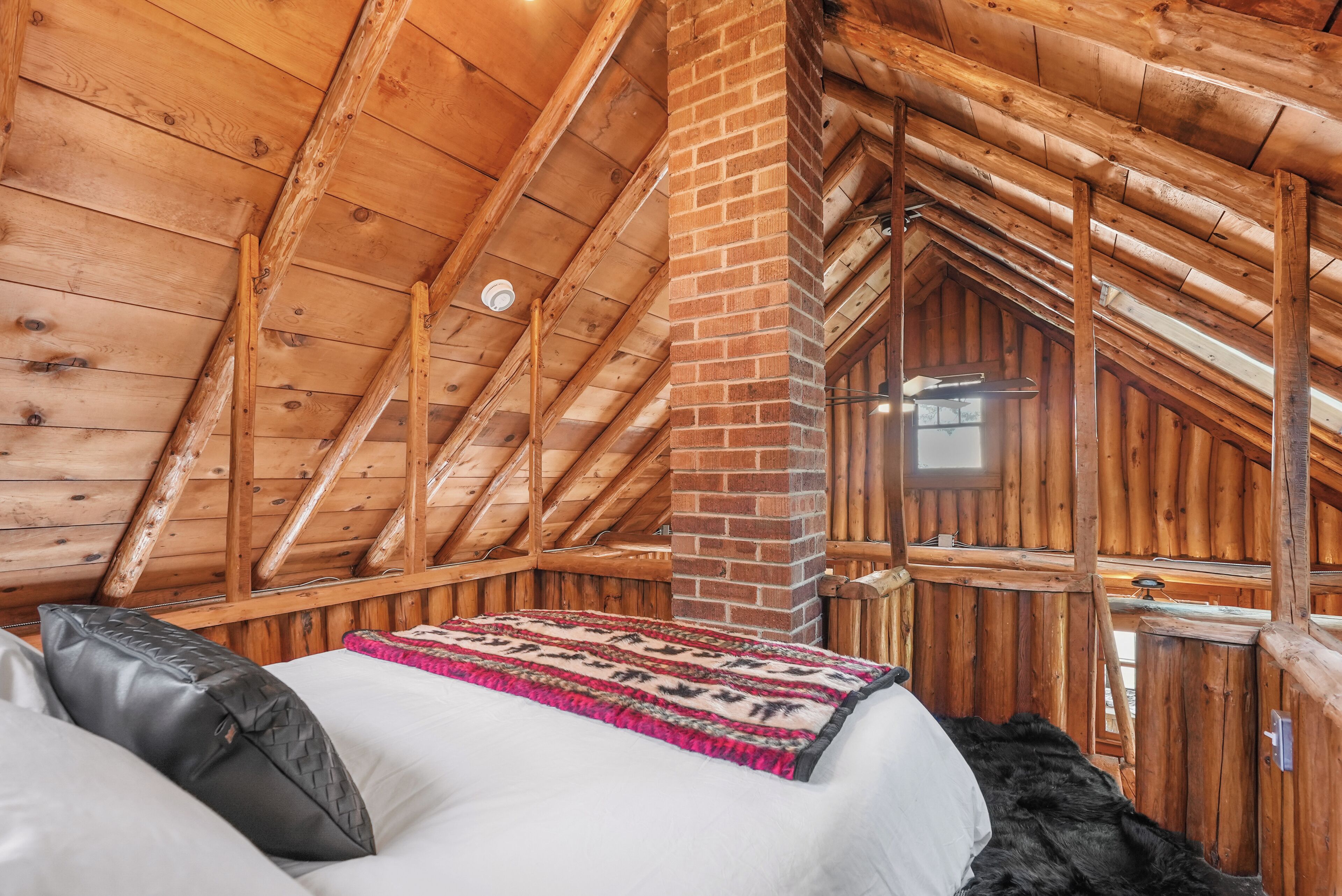 Une chambre confortable située dans les combles, avec des poutres en bois, une cheminée en briques et un lit recouvert d’une couverture à motifs.