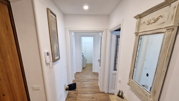 Un couloir avec un plancher en bois, des murs blancs et une porte en bois sur la gauche. Une petite table, ornée d’objets décoratifs, et un miroir se trouvent sur la droite.