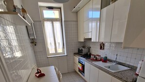 Une cuisine de taille réduite avec des meubles blancs, un pare-feu en carrelage, une fenêtre avec des rideaux, une table à manger avec des chaises et un four encastré.