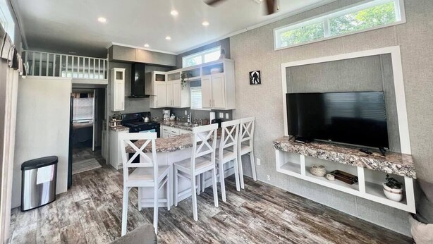 Una cucina moderna con isola centrale, mobili bianchi, una ventola a soffitto e un grande televisore montato a parete.