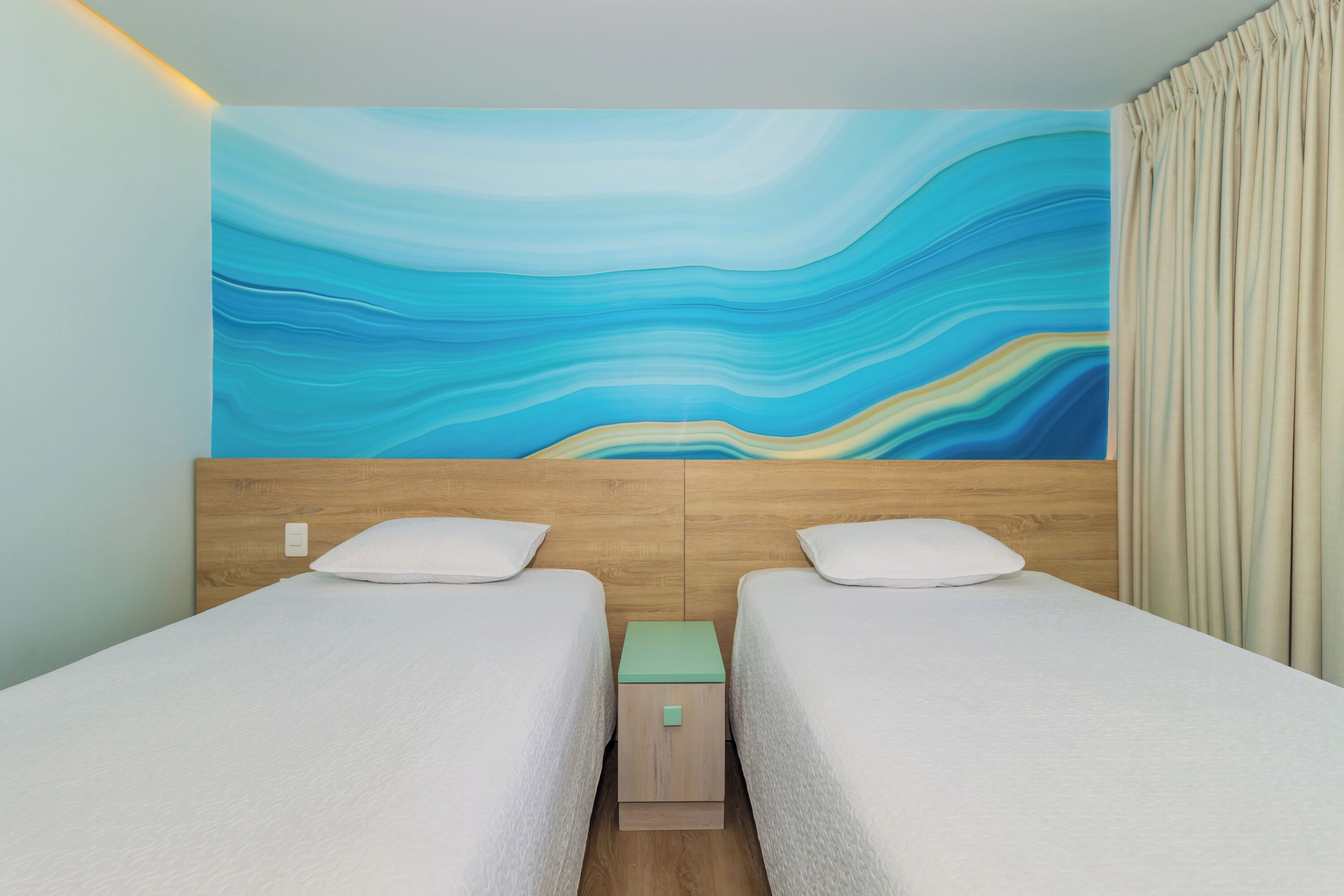 Quarto de hotel com duas camas, uma cabeceira de madeira, uma mesinha de cabeceira verde e um grande mural abstrato na parede.