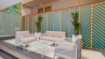Un espace de détente extérieur moderne, avec un canapé beige, des chaises blanches et une table basse en verre. On y trouve deux plantes en pot et un mur décoratif orné d’un motif géométrique.
