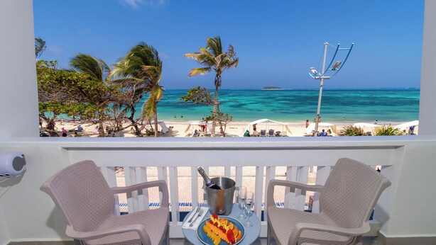 Un balcon avec deux chaises, une table sur laquelle se trouve une assiette de fruits et une bouteille de champagne, et une vue sur la plage et l’océan.
