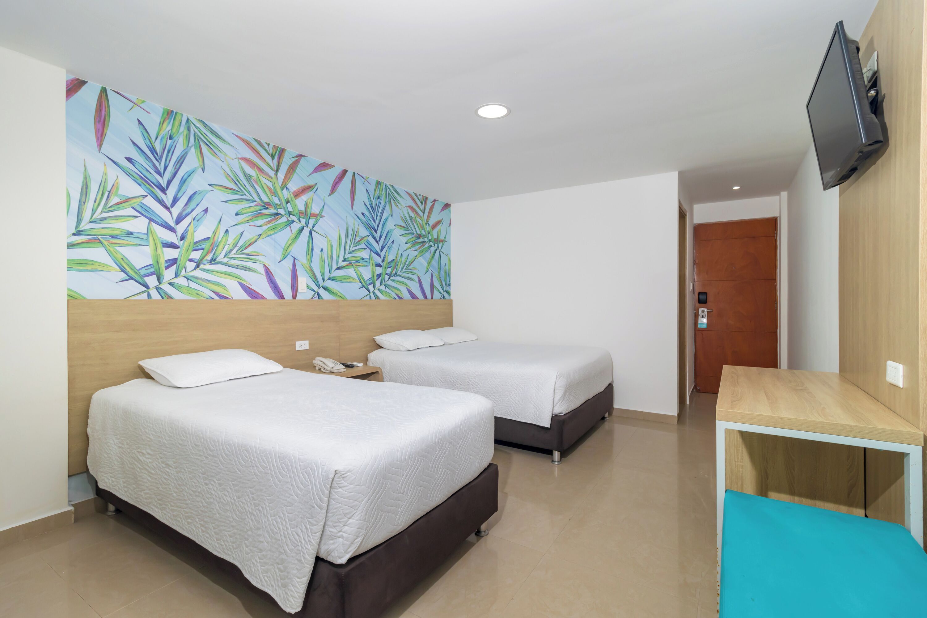 Quarto de hotel com duas camas, cabeceira de madeira, painel na parede, televisão fixada na parede e uma mesinha.