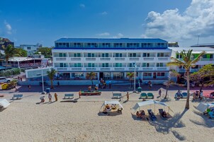 Un hotel de varios pisos con balcones, ubicación frente a la playa y una zona de arena con tumbonas.