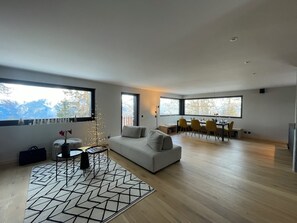 Ein modernes Wohnzimmer mit einer Couch, einem Essbereich mit Stühlen und großen Fenstern mit Blick ins Freie.