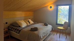 Una camera da letto con soffitto spiovente, un letto con coperta a scacchi, una sedia e una finestra vista alberi.